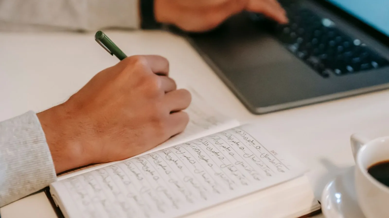 Writing in a notebook beside a laptop and coffee